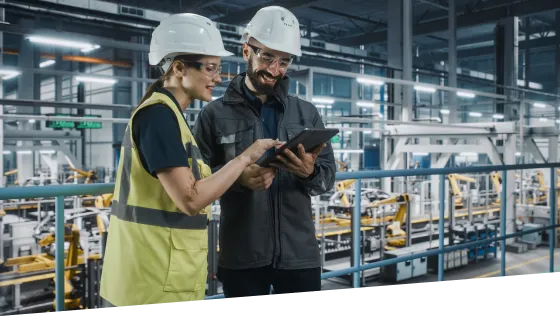 A man and a woman dressed in high visibility vests, using a tablet. It looks like they are in a manufacturing facility. 